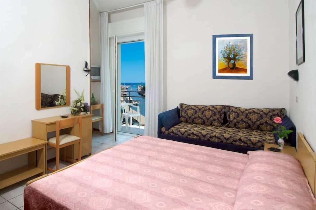 A hotel room with a bed, a desk, a chair, a sofa, a painting, and a view of the sea.