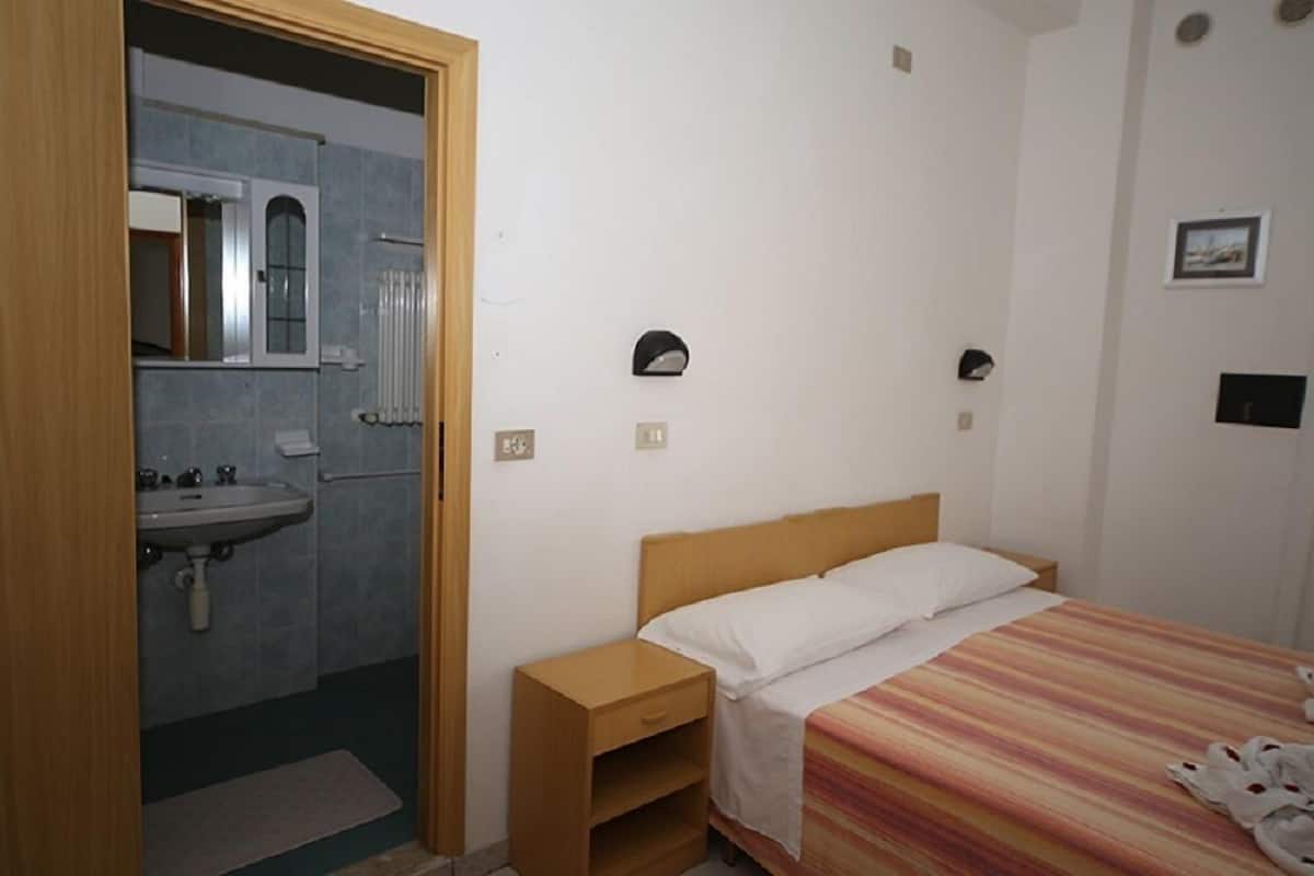 A bedroom with a single bed, a wooden nightstand, a bedside table, a wall-mounted light, and a bathroom with a sink and mirror.