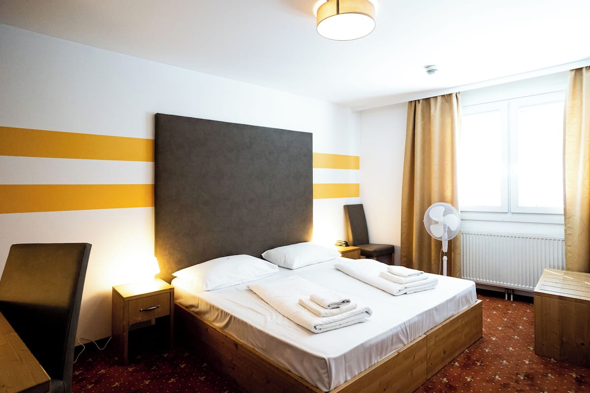 A hotel room with a large bed, bedside tables, a desk, a chair, a standing fan, and a window with curtains.