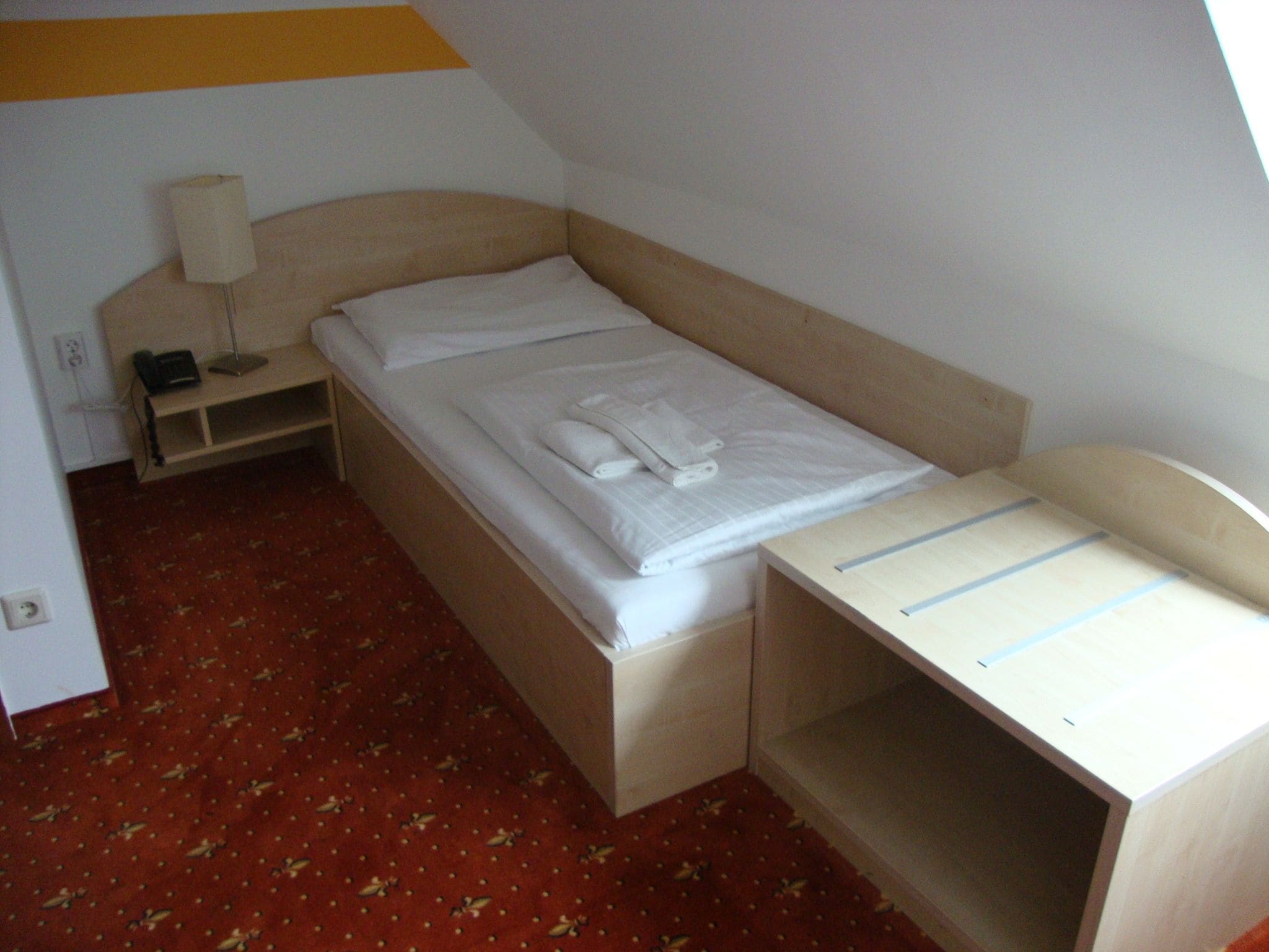 A small, single-bed room with a wooden headboard, a bedside table with a lamp, and a red carpet with a leaf pattern.