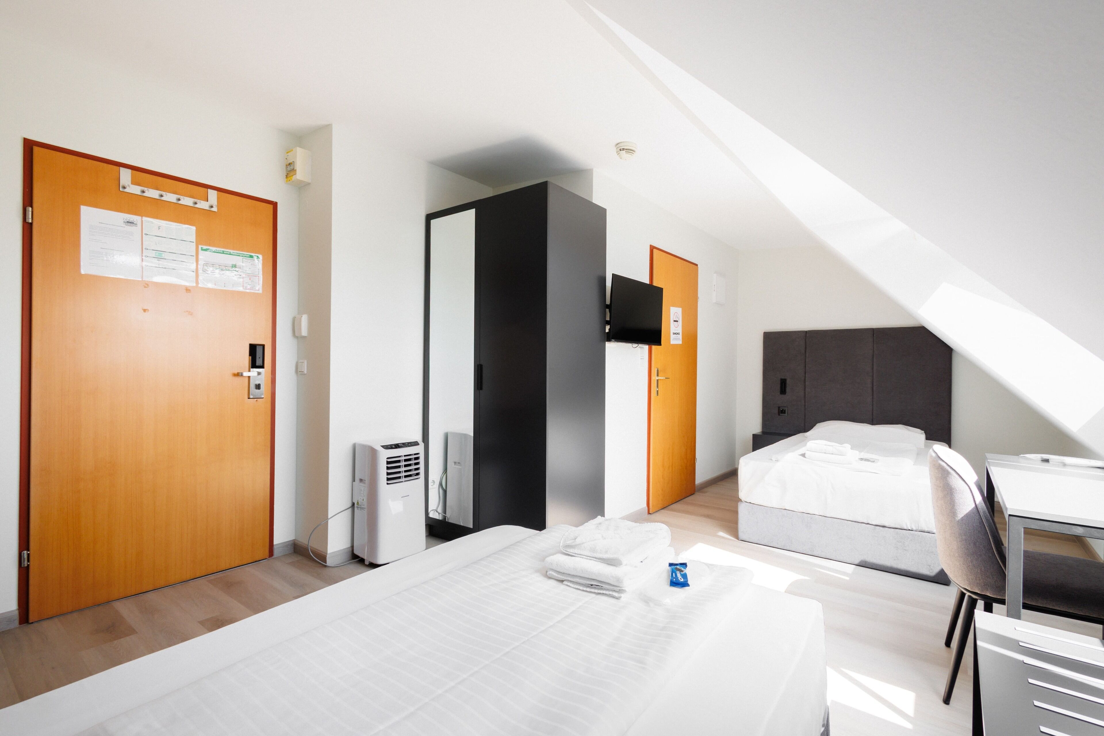 Habitación de hotel con cama, escritorio y silla. Se observa una puerta con un letrero, televisor montado en la pared y un termostato.