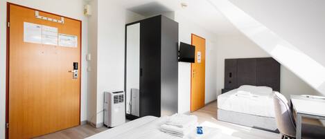Quarto de hotel com cama, escrivaninha e cadeira. Há uma porta com placa, uma TV fixada na parede e um termostato.