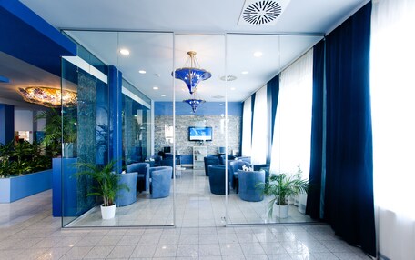A modern lobby with glass walls, blue seating, potted plants, and a chandelier.