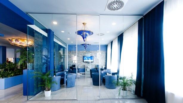 A modern lobby with glass walls, blue seating, potted plants, and a chandelier.