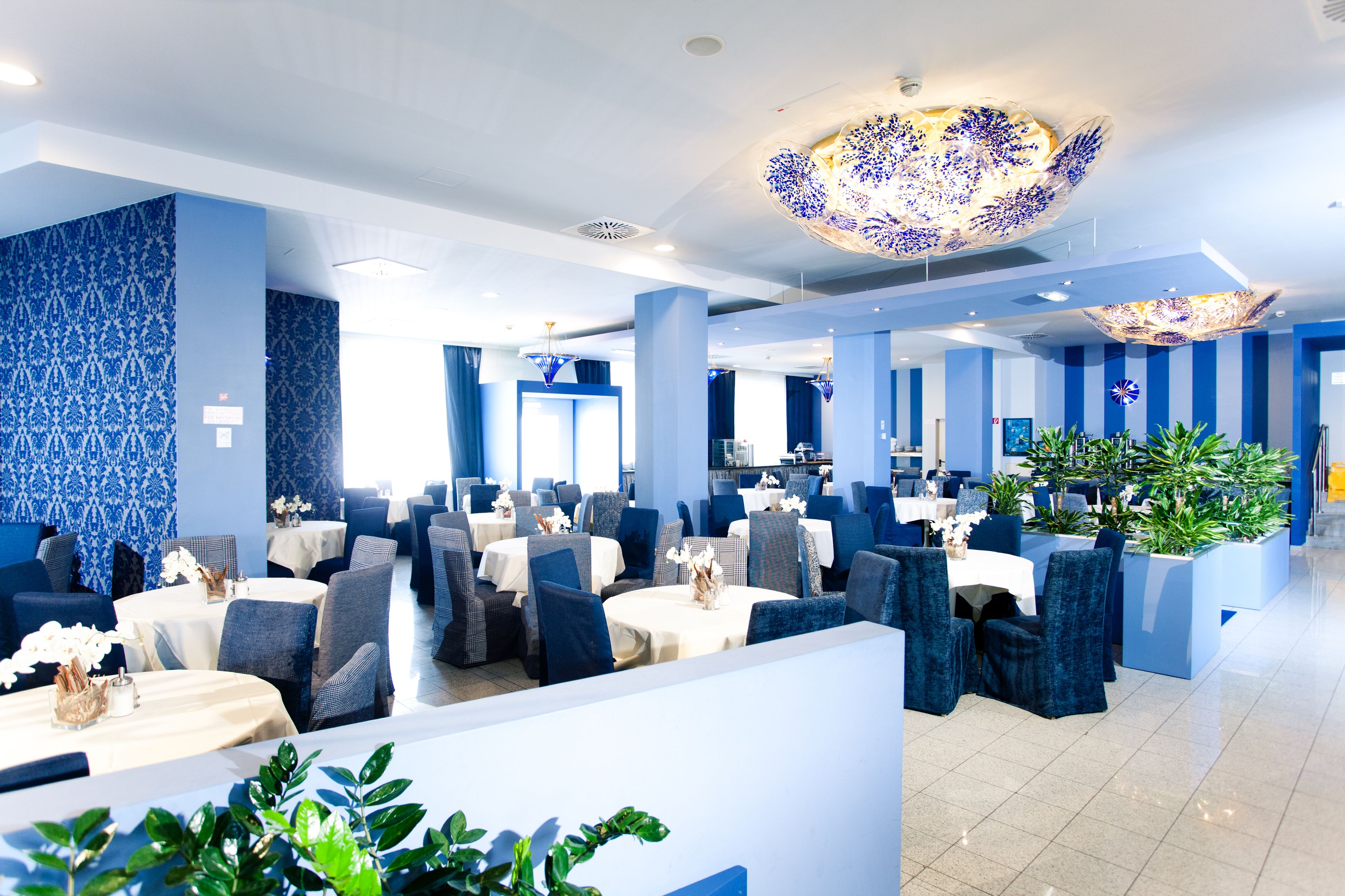 Un restaurante moderno con decoración azul y blanca, varias zonas de comedor y plantas en macetas.