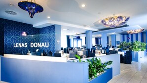 A modern hotel lobby with a reception desk labeled "LENAS DONAU," featuring a blue and white color scheme, contemporary lighting fixtures, and indoor plants.