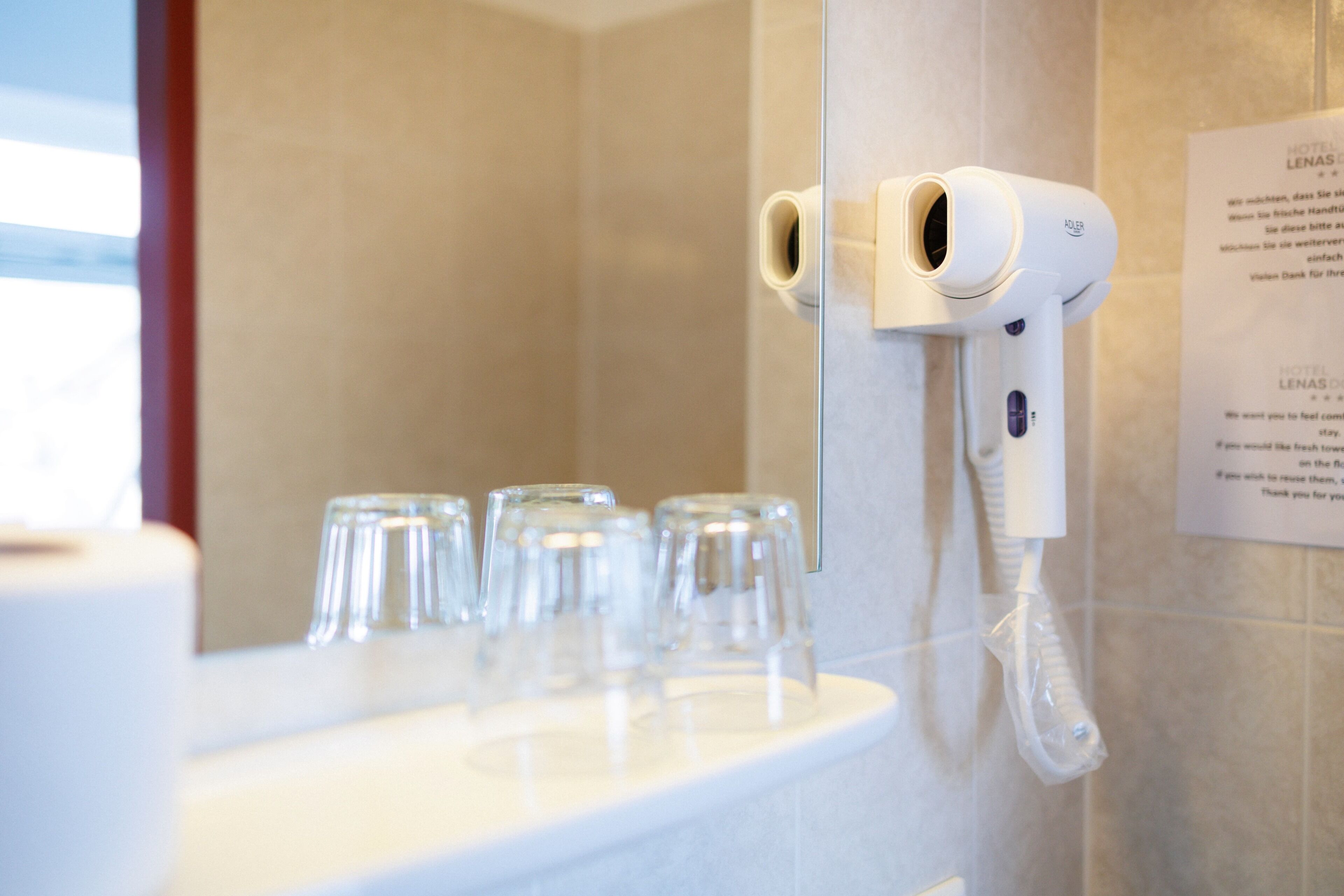 Een hotelbadkamer met een haardroger aan de wand, een spiegel en een aanrecht met glazen.