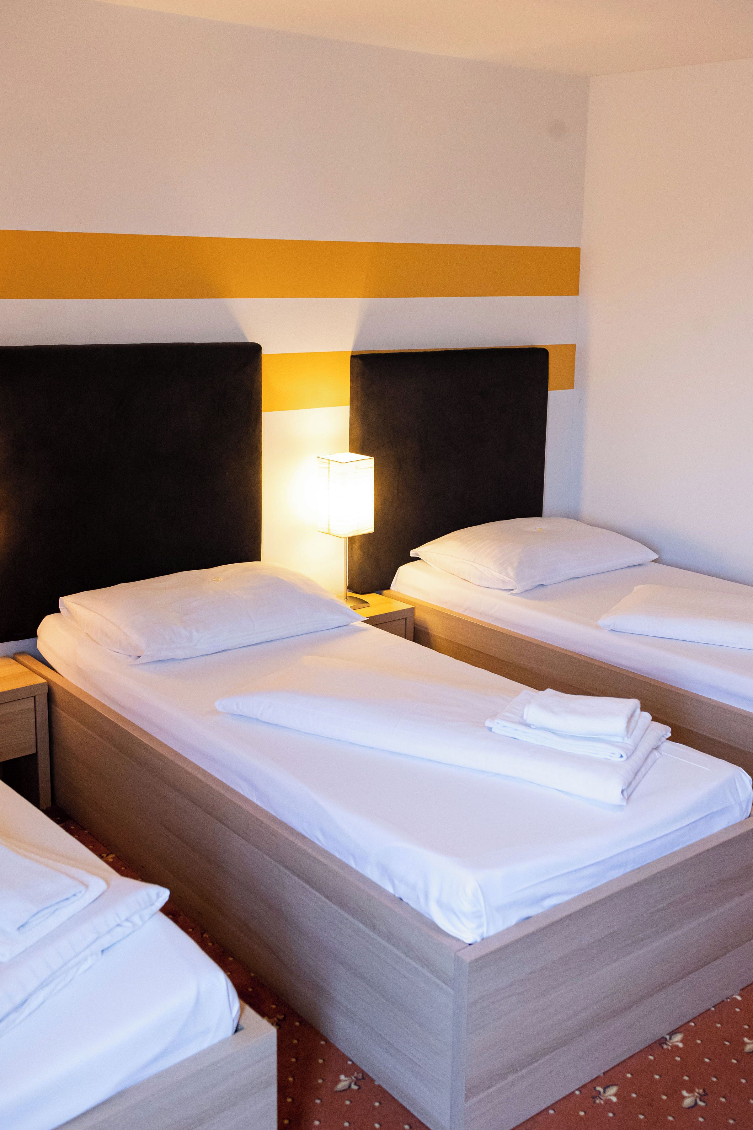 A hotel room with two single beds, each with white linens and pillows, a bedside table with a lamp, and a wooden headboard.