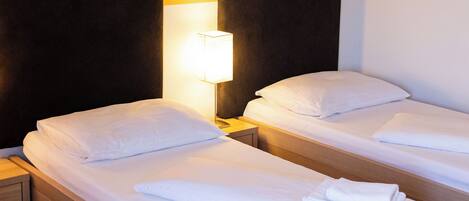 A hotel room with two single beds, each with white linens and pillows, a bedside table with a lamp, and a wooden headboard.