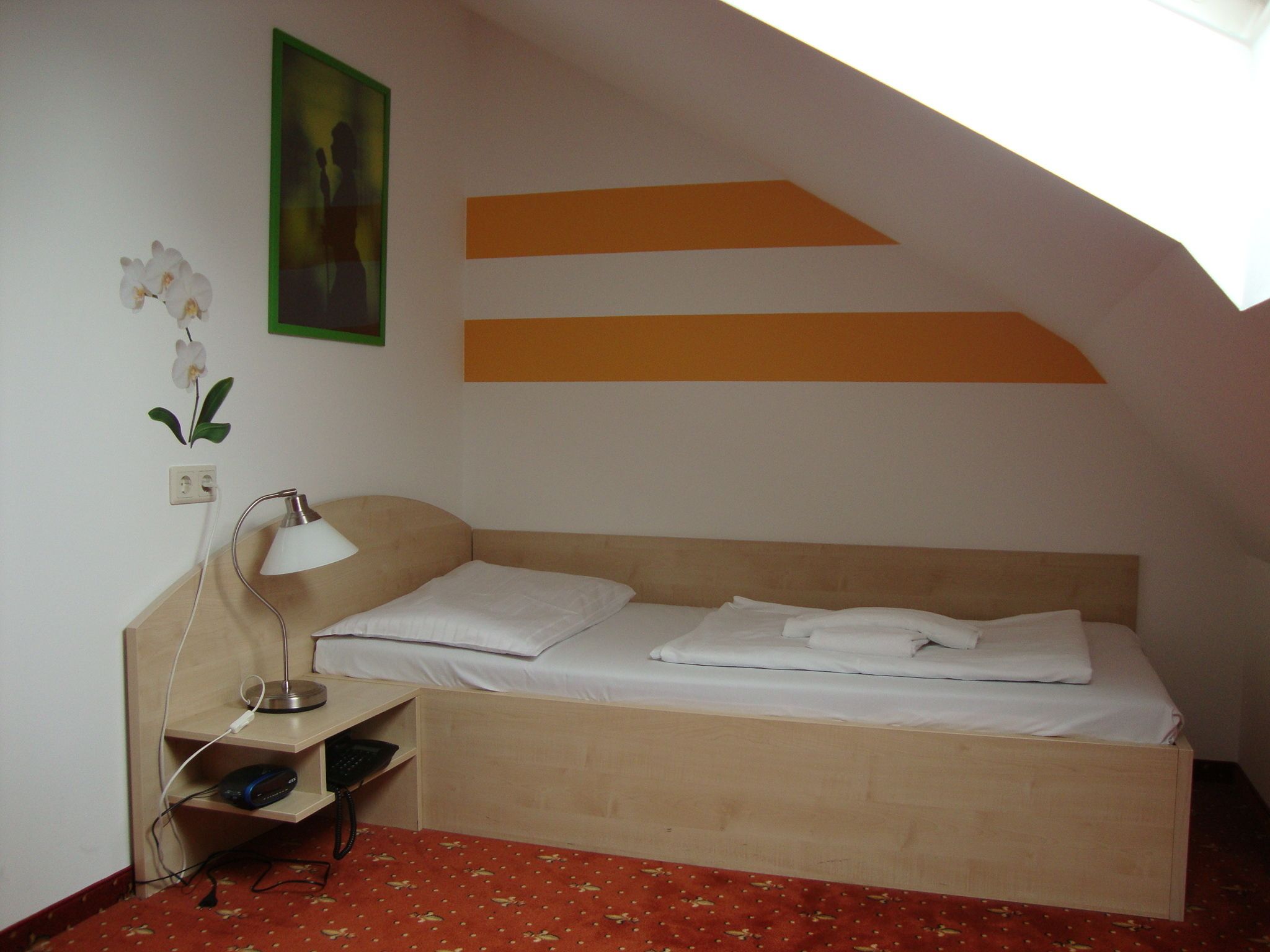 Ein kleines Zimmer mit einem Einzelbett, geneigter Decke, Nachttisch mit Lampe, Steckdose und kleinem Ablagefach.