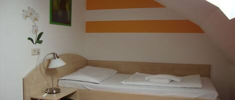 Una habitación pequeña con cama individual, techo inclinado, mesita de noche con lámpara, toma de corriente y un estante pequeño.