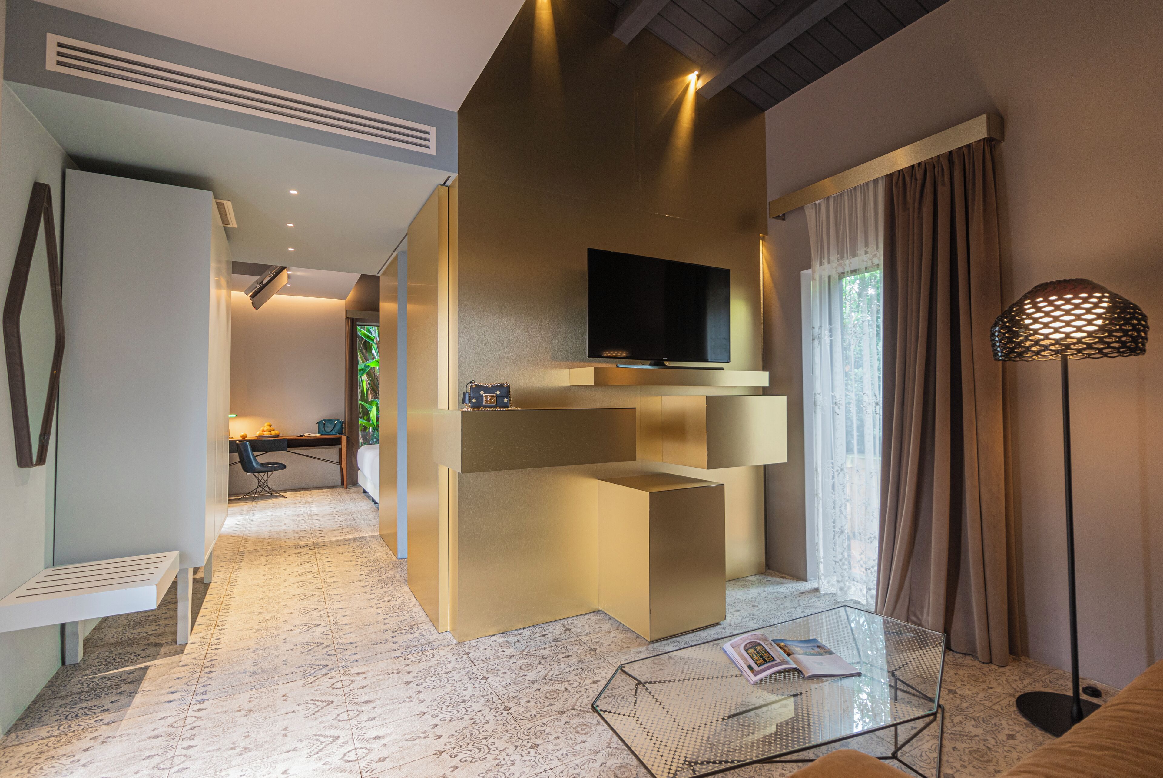 Una habitación de hotel moderna con un televisor de pantalla plana montado en la pared, una mesa de centro de vidrio y una zona de estar con un banco y un escritorio.
