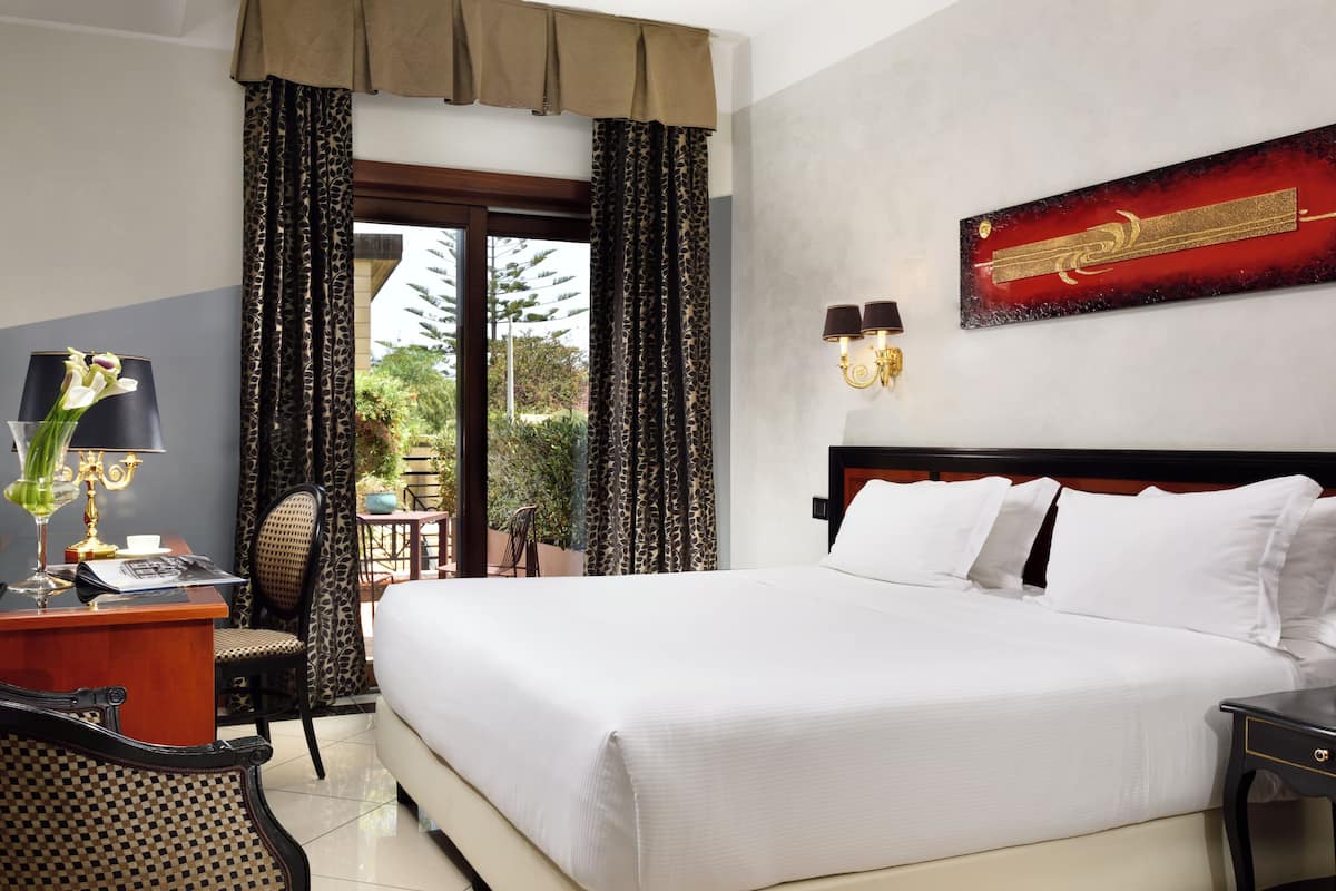 A hotel room with a large bed, a desk with a lamp, a chair, and a view of the outdoors.