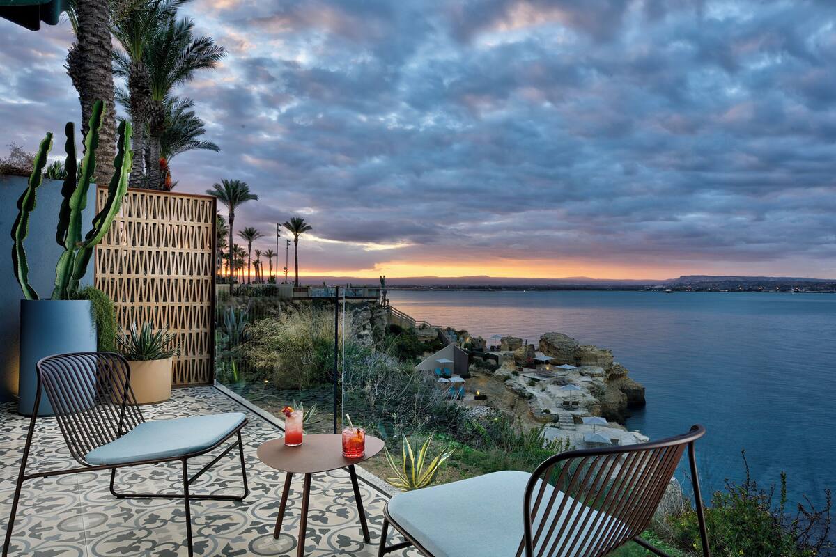 A patio with two chairs and a table, overlooking a coastal landscape with cacti and a view of the sea at sunset.