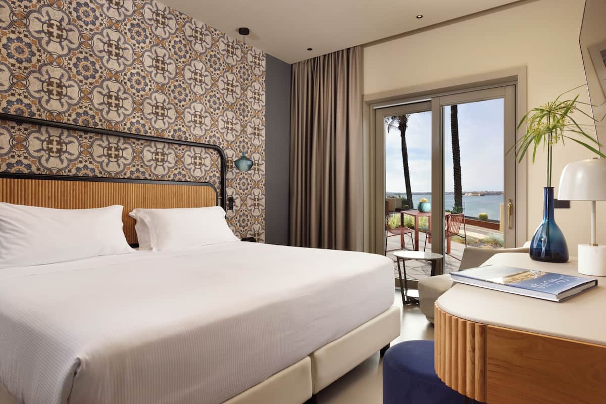A hotel room with a large bed, a desk with a lamp, and a view of the sea.