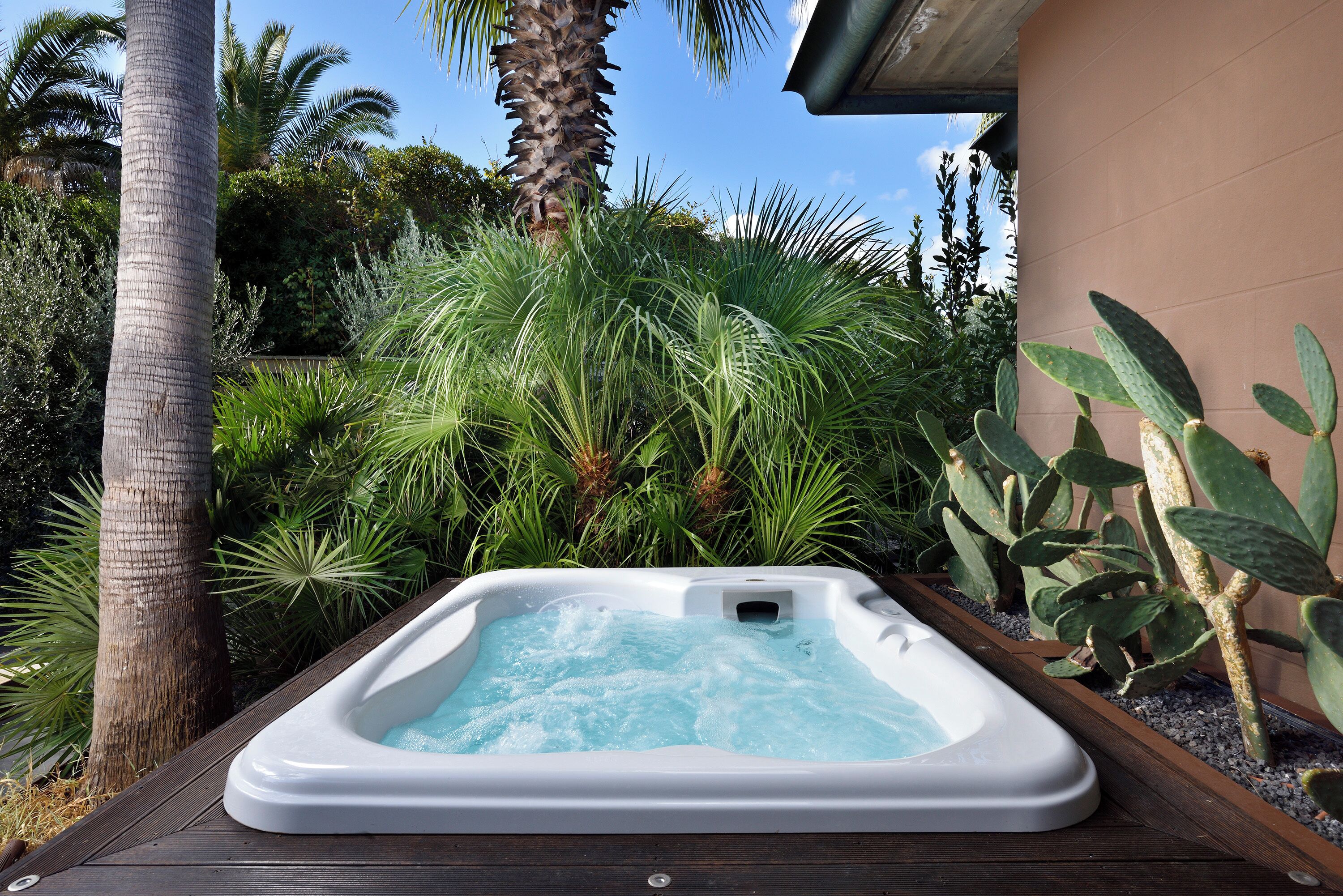 A hot tub surrounded by lush greenery, including palm trees and various plants, set on a wooden deck.