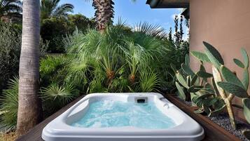 A hot tub surrounded by lush greenery, including palm trees and various plants, set on a wooden deck.