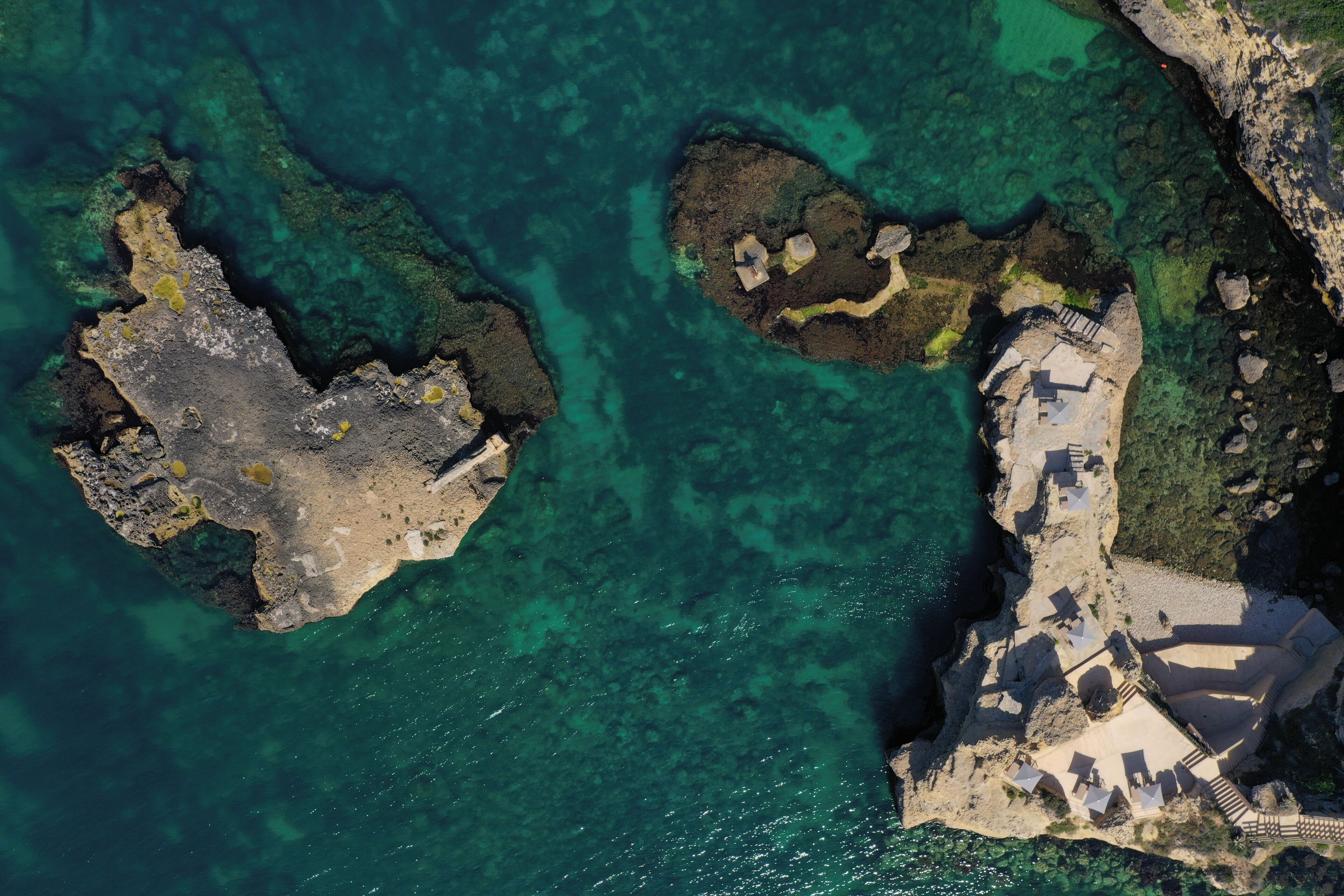 An aerial view of a coastal area with clear turquoise waters, rocky formations, and a small building with steps leading to a beach area.