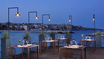 A waterfront restaurant with tables set for dining, overlooking a cityscape and the sea.