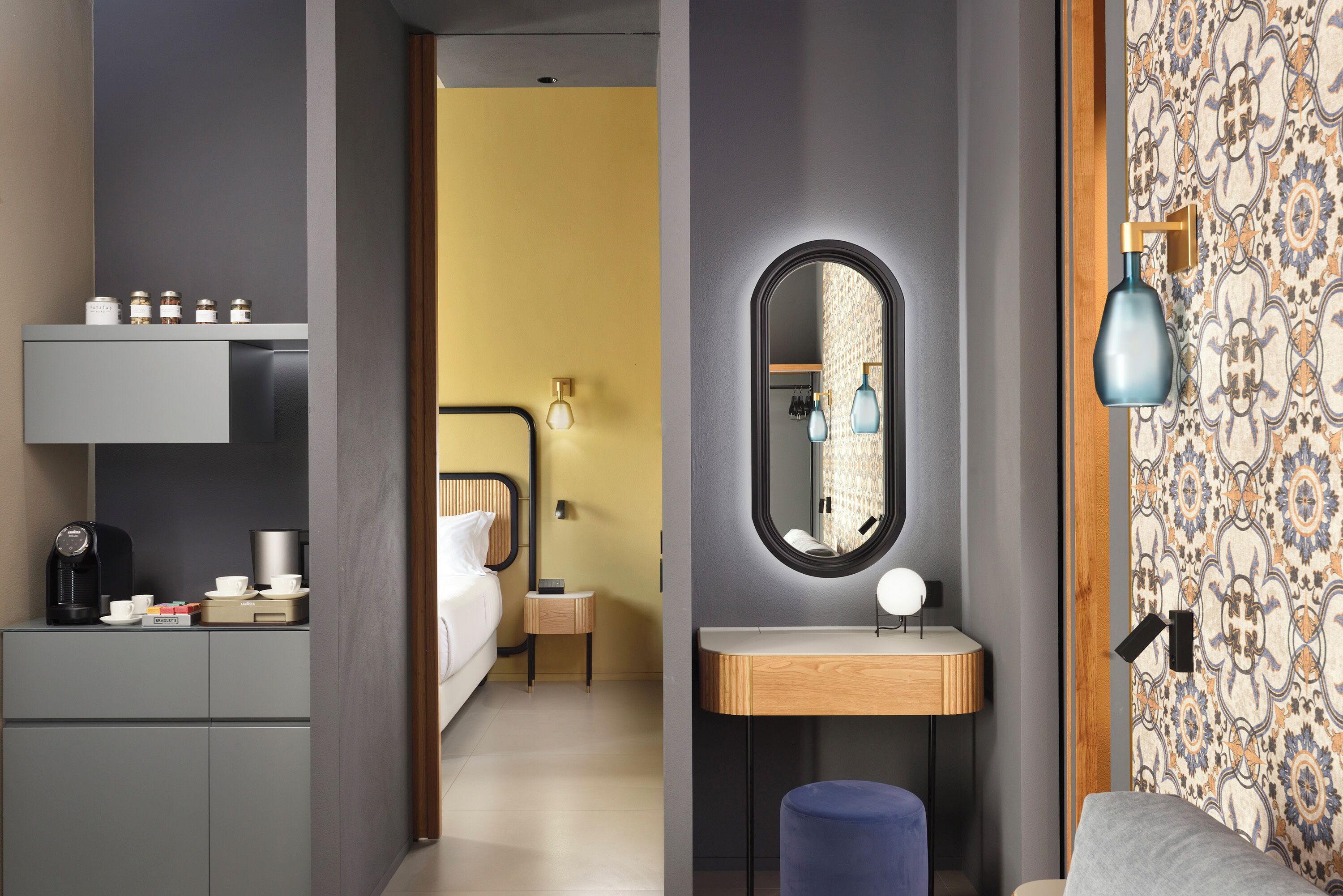 Ein Hotelzimmer mit Bett, Nachttisch und Spiegel. Außerdem ein kleiner Hocker und eine Wandlampe.