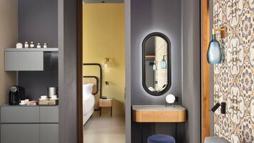 Ein Hotelzimmer mit Bett, Nachttisch und Spiegel. Außerdem ein kleiner Hocker und eine Wandlampe.
