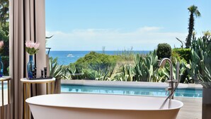 A modern bathroom with a freestanding bathtub, a small table with toiletries, and a view of the ocean.