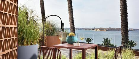 A terrace with a table and chairs, overlooking a body of water and a distant cityscape.