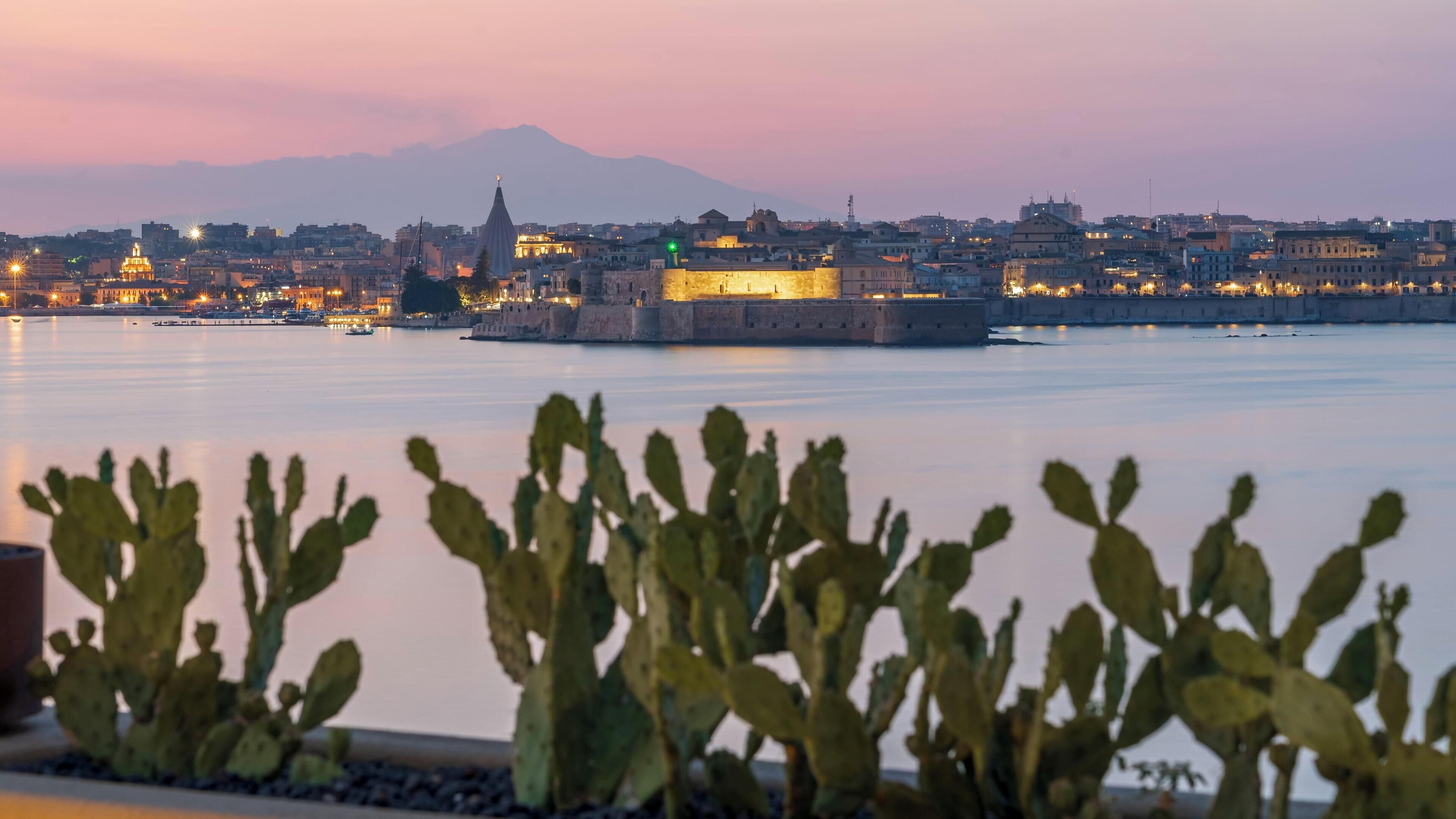 A cityscape at dusk with a prominent building and a body of water in the foreground.