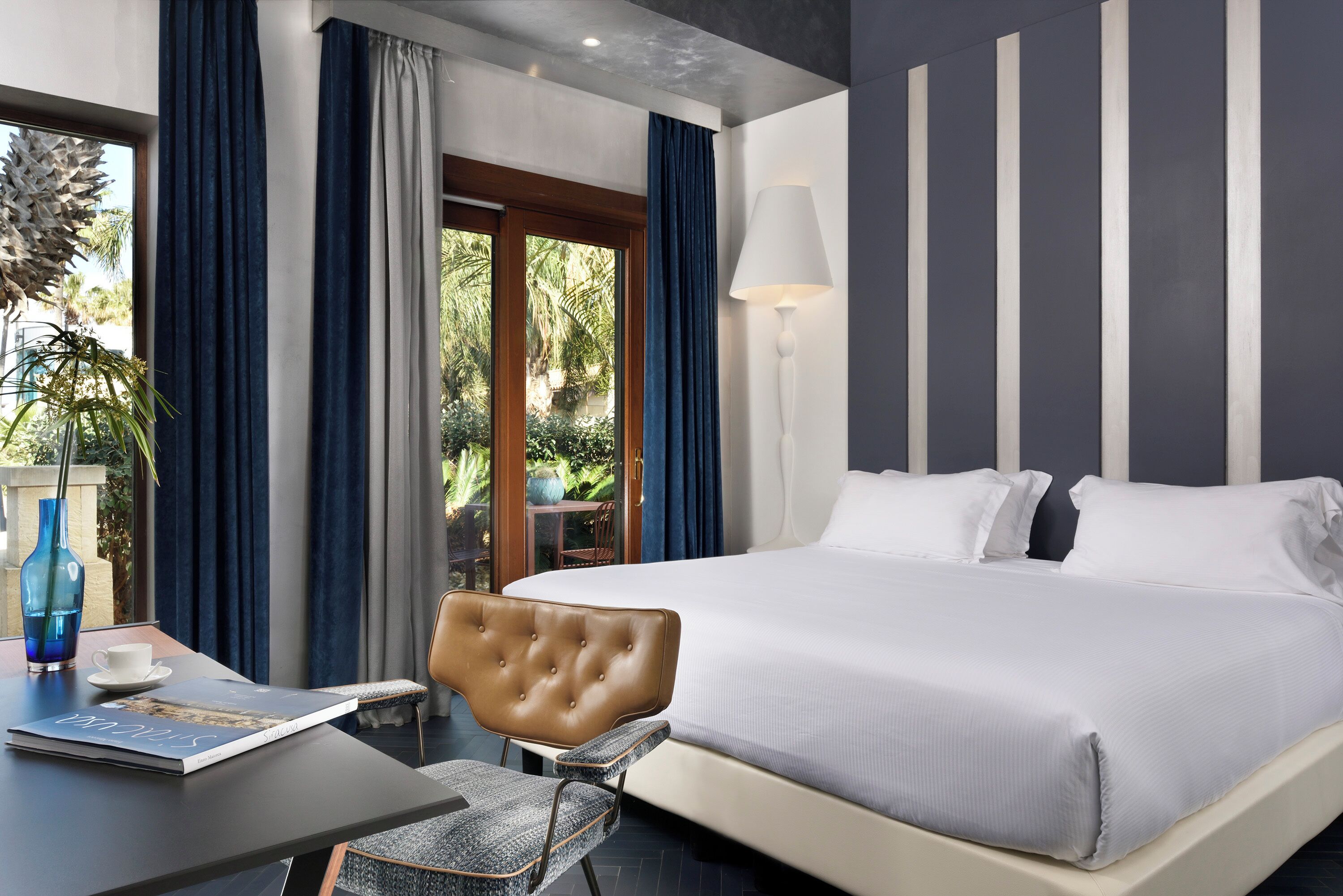 A hotel room with a large bed, a desk with a chair, a lamp, and a view of greenery outside.