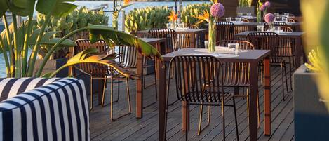 An outdoor dining area with wooden tables and chairs, surrounded by potted plants and a view of the sea.