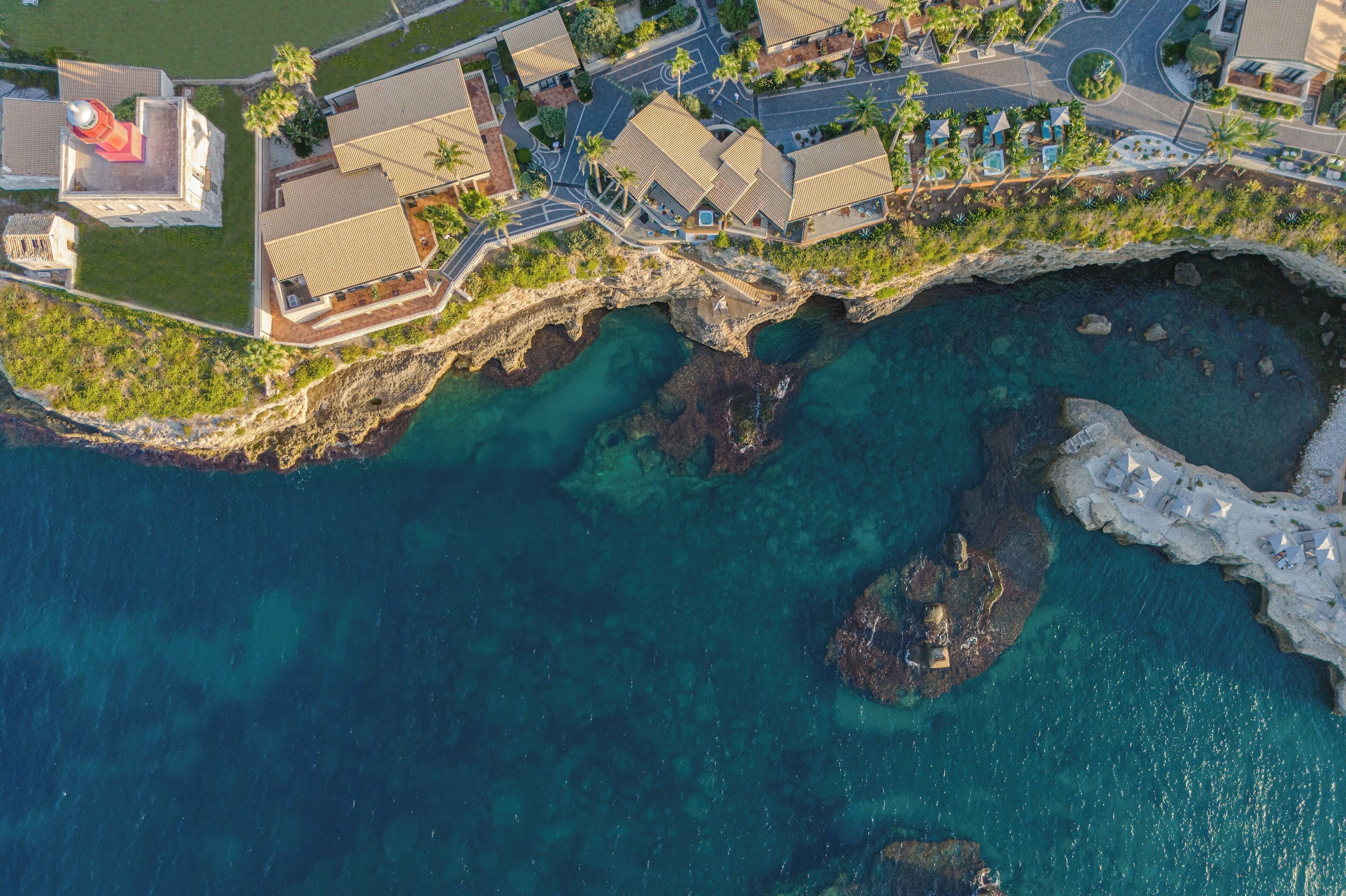 An aerial view of a coastal hotel with multiple buildings, a rocky shoreline, and clear blue waters.