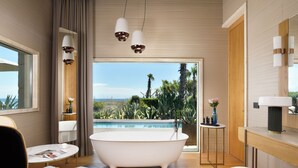 A modern bathroom with a freestanding bathtub, a round table with a magazine, and a view of a pool and greenery.
