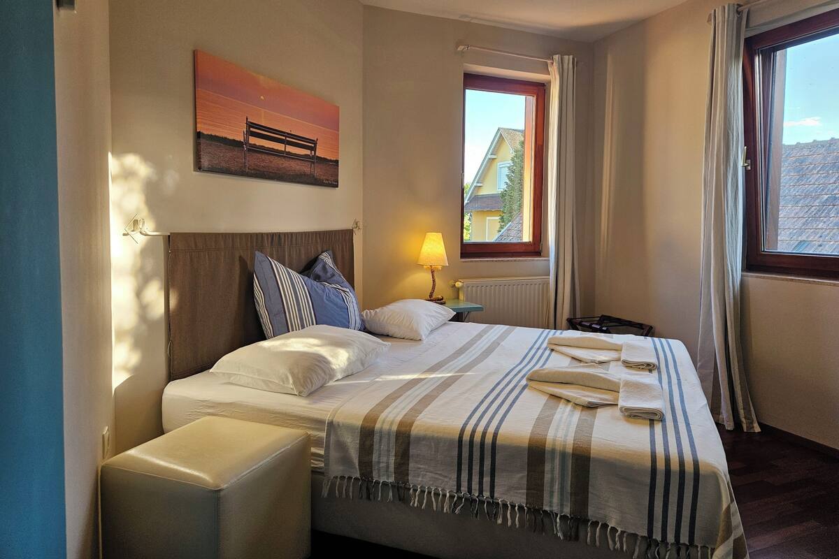 A hotel room with a bed, a window with curtains, a nightstand, a lamp, and a painting on the wall.