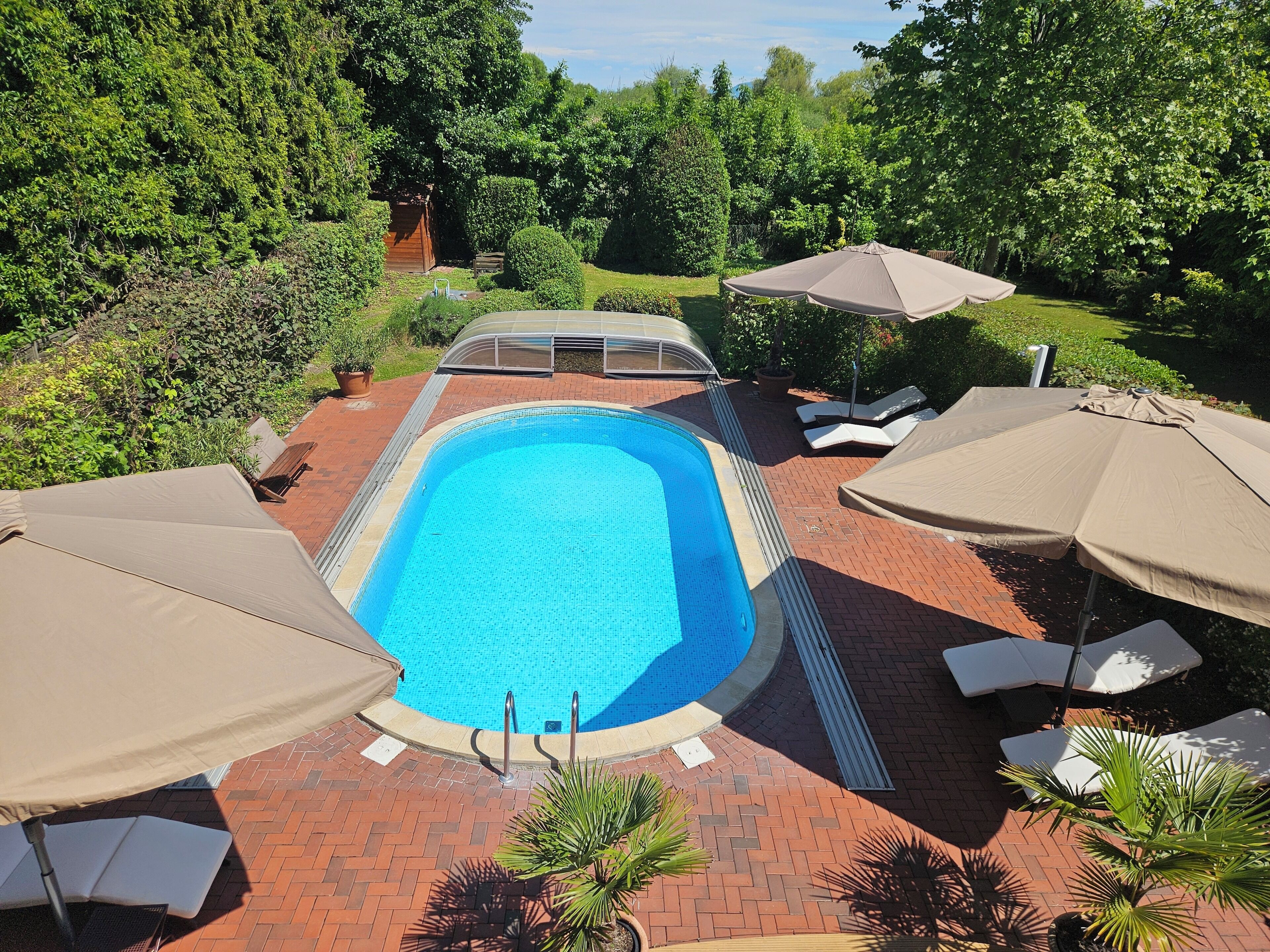 A rectangular swimming pool with a tiled edge, surrounded by a brick patio. There are lounge chairs and umbrellas around the pool area.