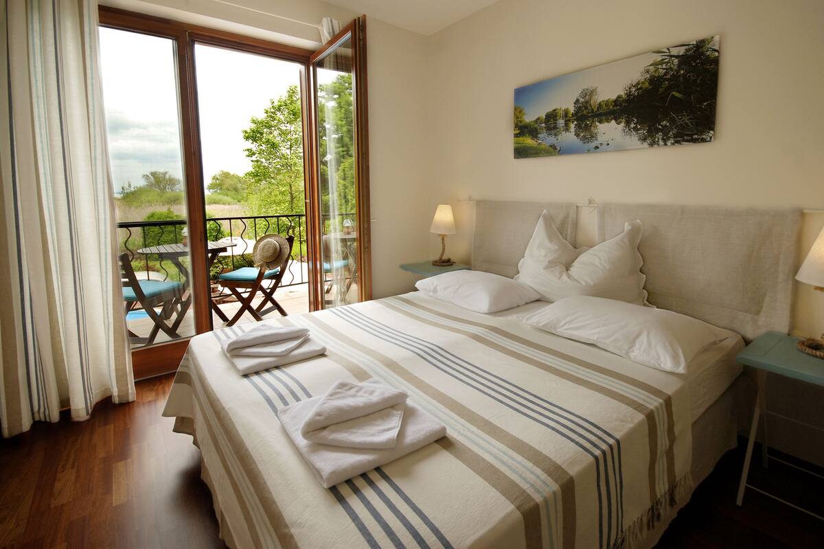 A bedroom with a large bed, a view of the outdoors, a painting on the wall, and a balcony with a table and chairs.