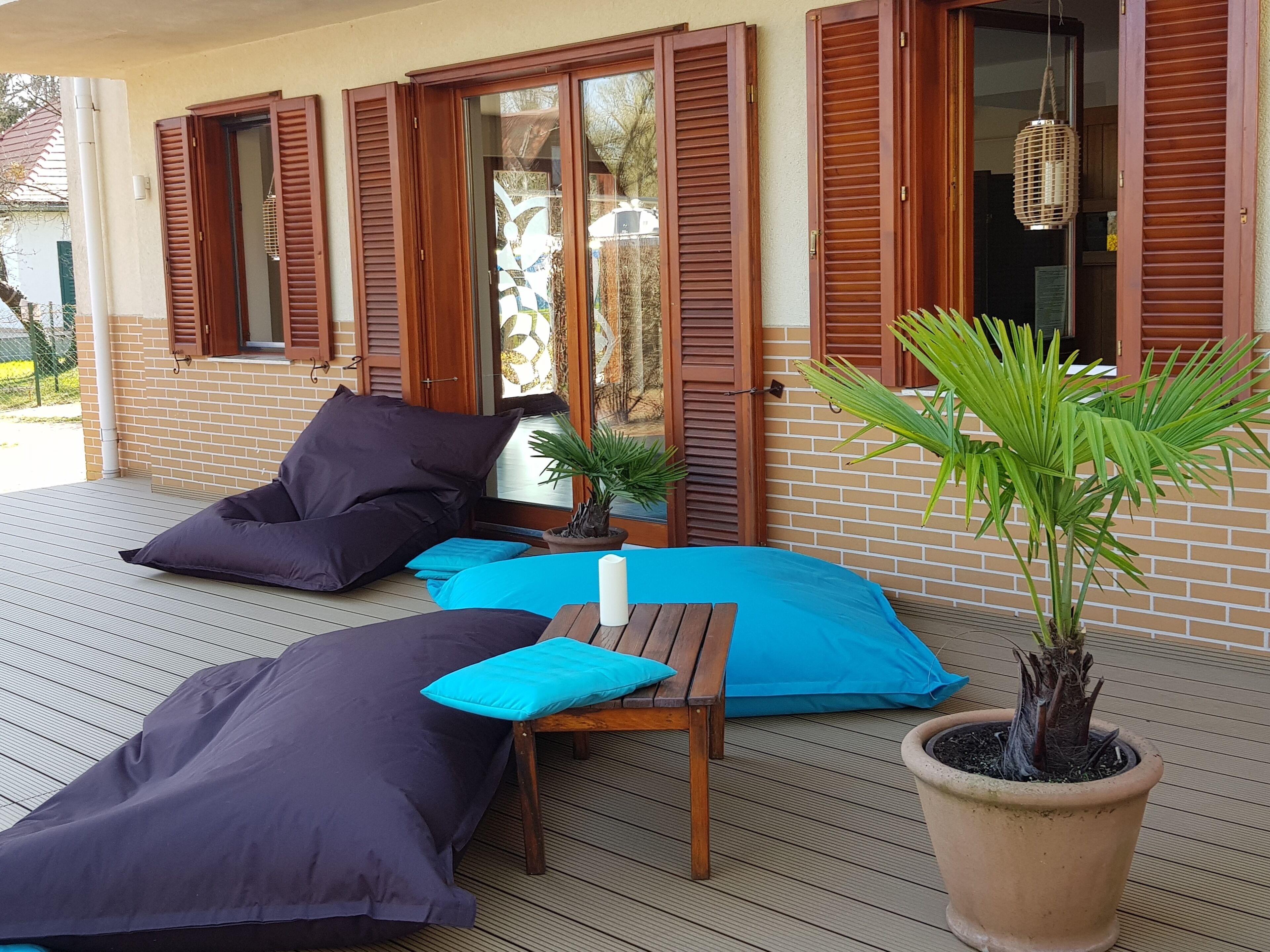 A covered outdoor seating area with wooden furniture and cushions, a potted plant, and a brick wall with wooden shutters.