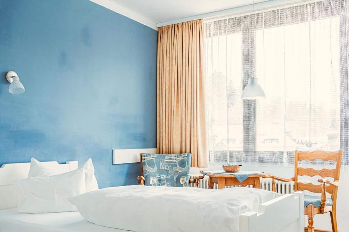 A hotel room with a large bed, a wooden chair, a small table with a bowl, and a window with curtains.
