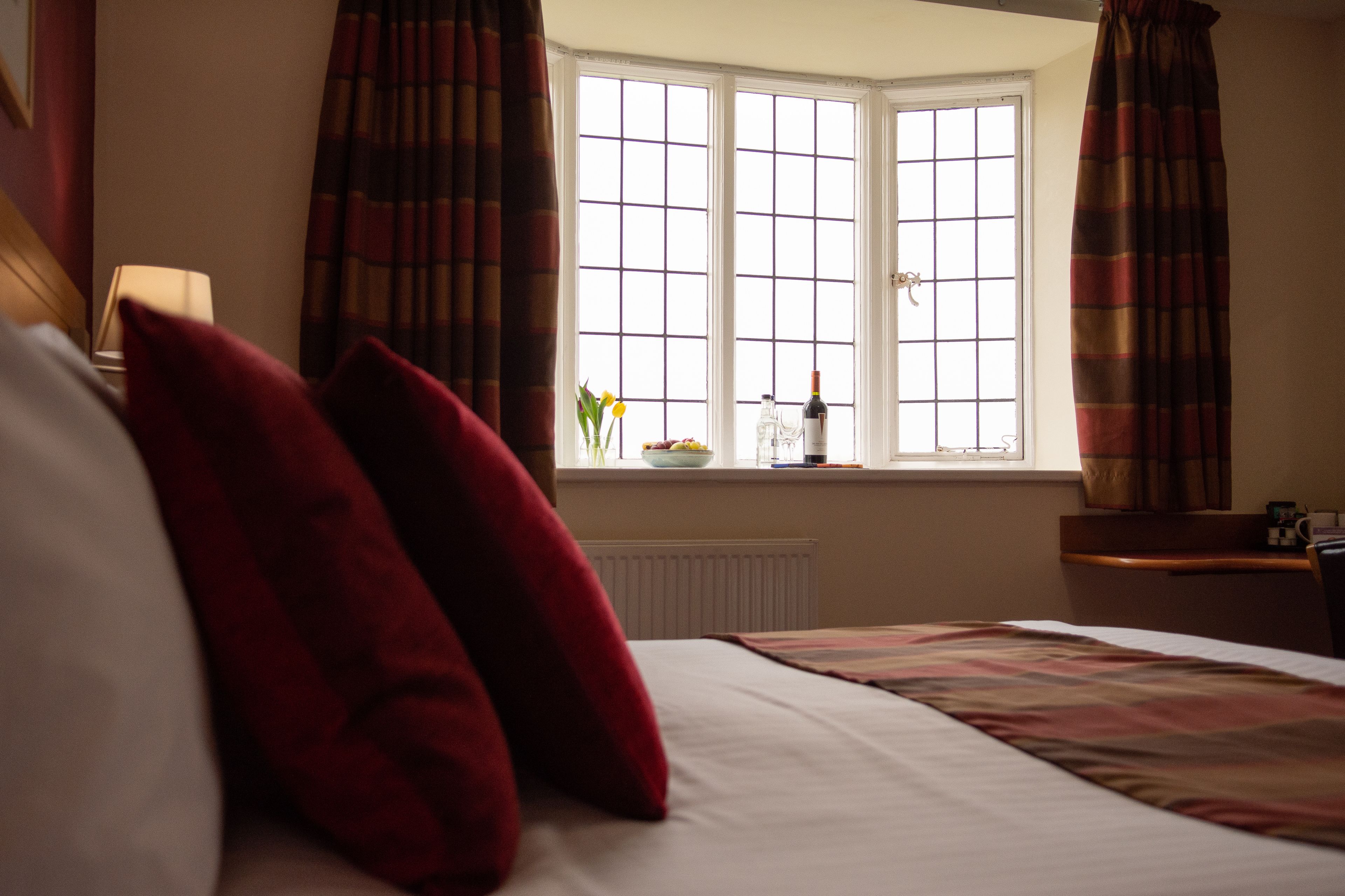 A hotel room with a bed, red pillows, a window with curtains, a vase with flowers, and a bottle on the windowsill.