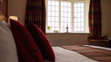 A hotel room with a bed, red pillows, a window with curtains, a vase with flowers, and a bottle on the windowsill.