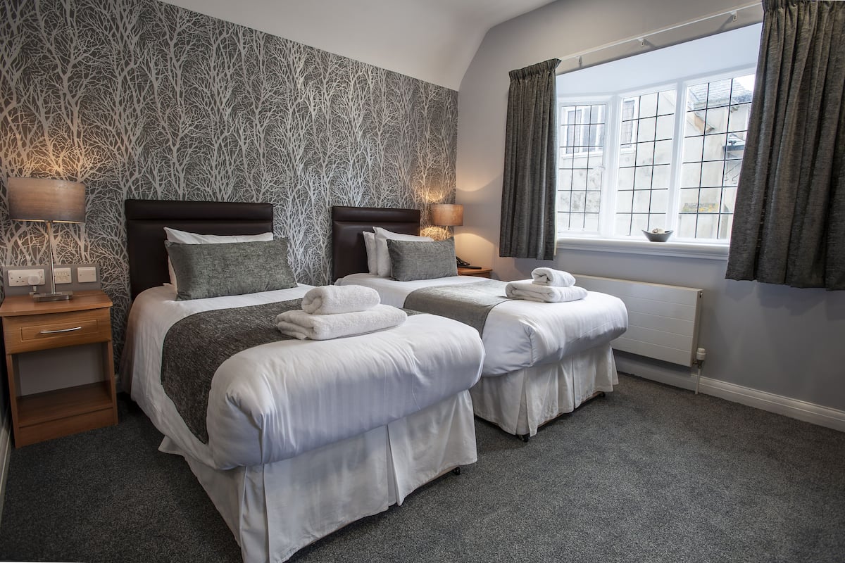 A hotel room with two beds, a window with curtains, a radiator, and a patterned wallpaper.