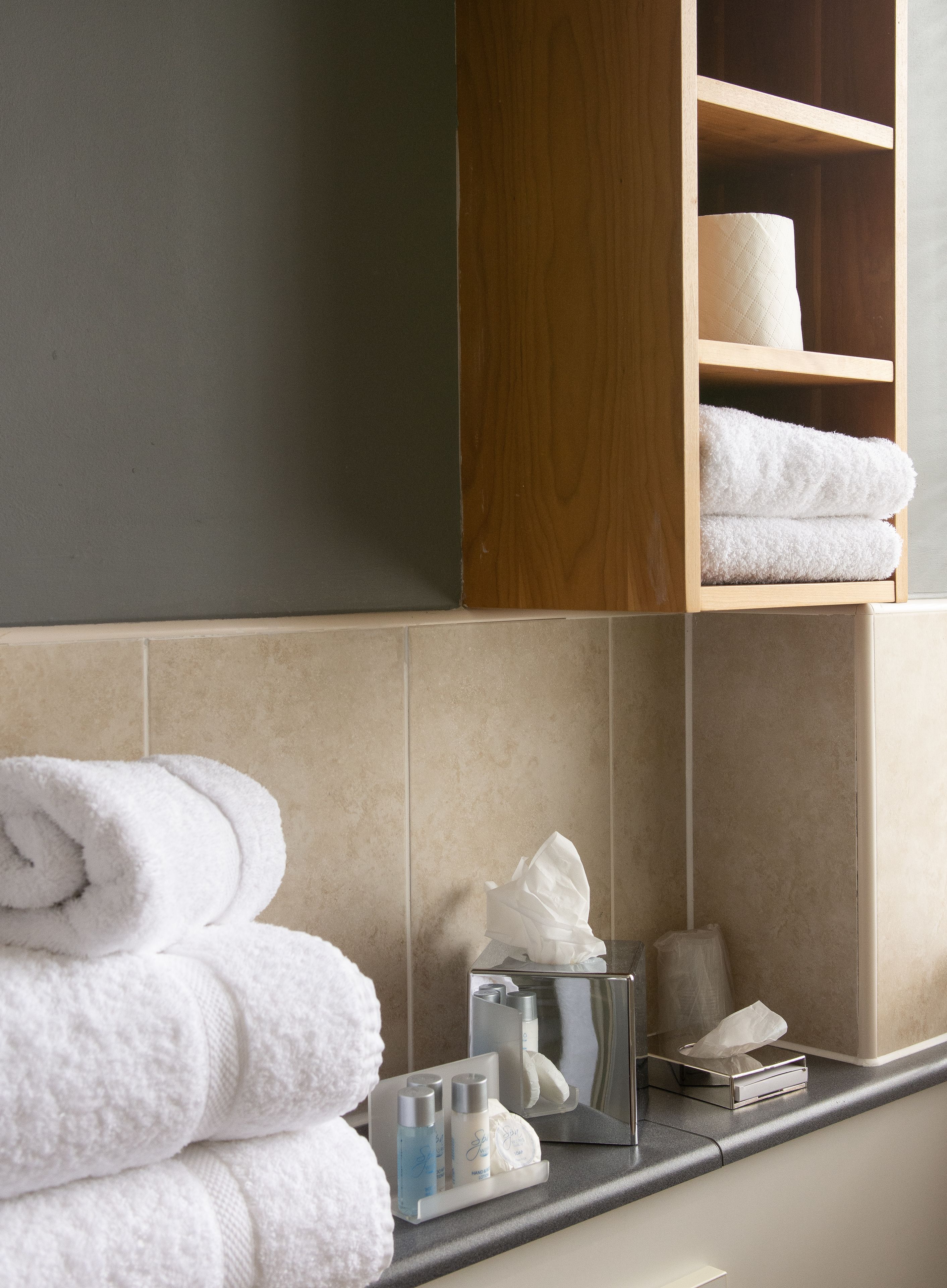 A bathroom with a wooden shelf holding rolled towels, a mirror, and a towel dispenser.