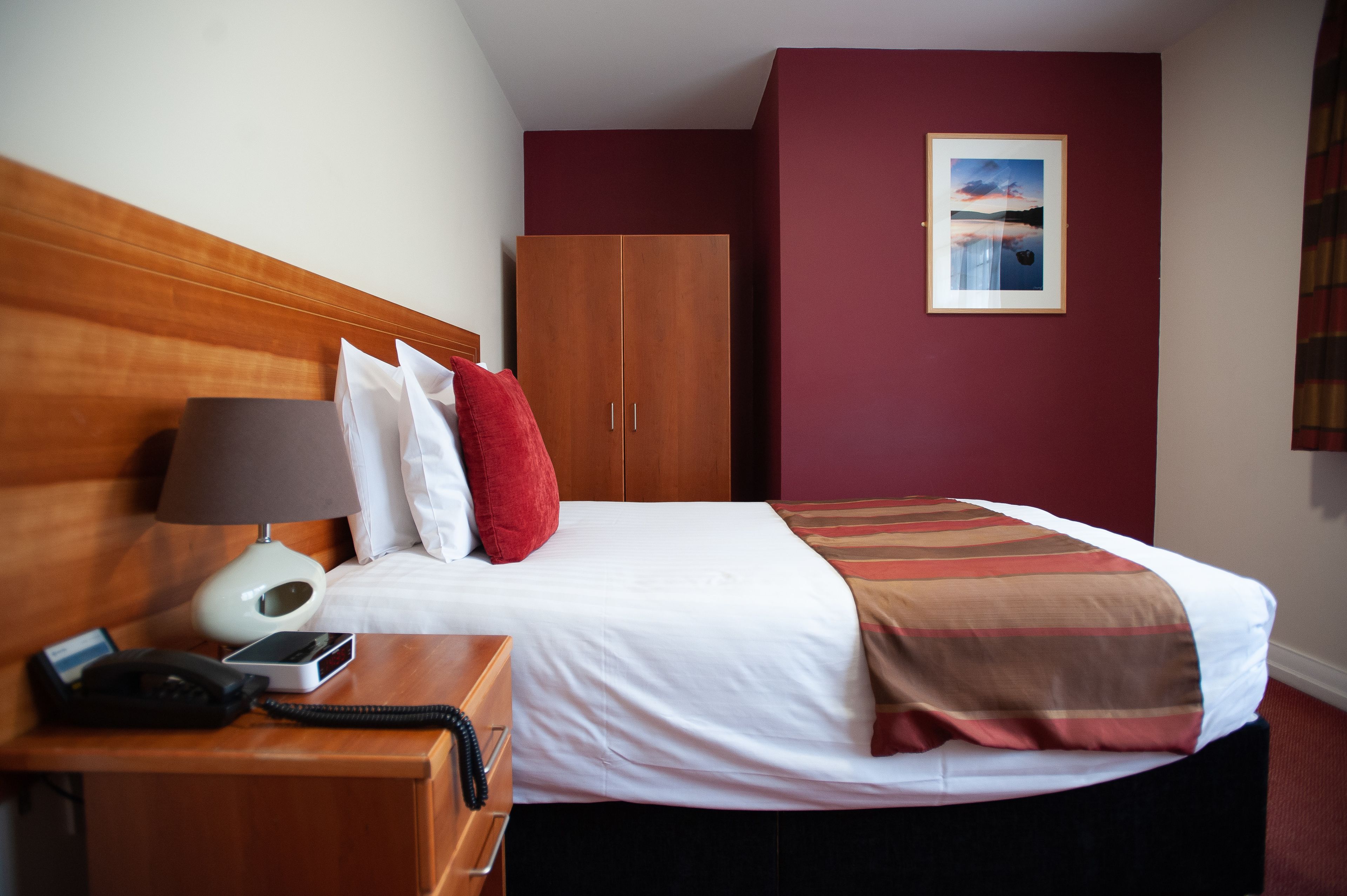 A hotel room with a wooden headboard, a bed with white linens, a red pillow, a brown and white striped blanket, a wooden wardrobe, a framed picture on the wall, and a bedside table with a lamp, phone, and a card.
