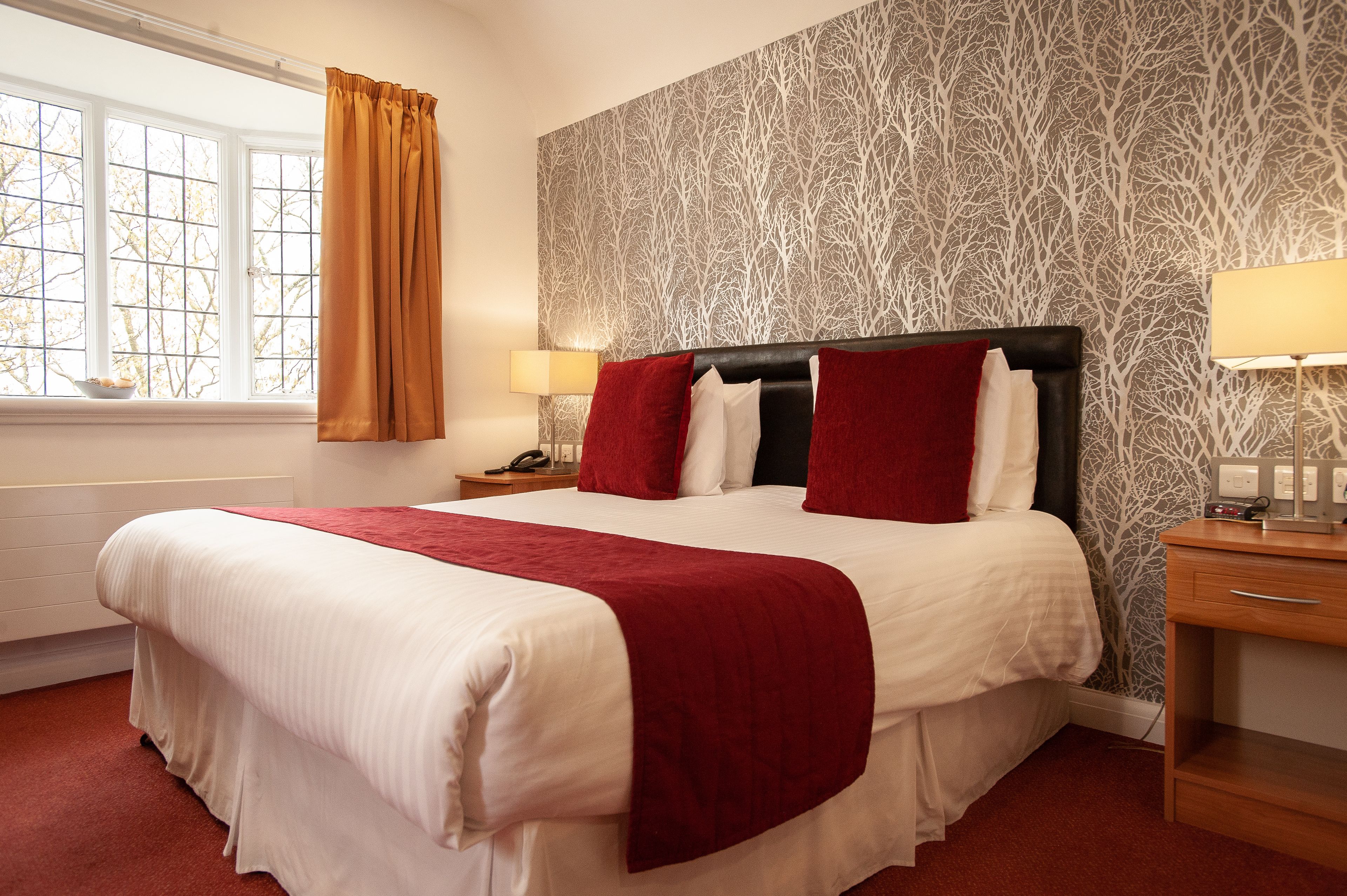 A hotel room with a large bed, a window with curtains, a patterned wallpaper, and a bedside table with a lamp.
