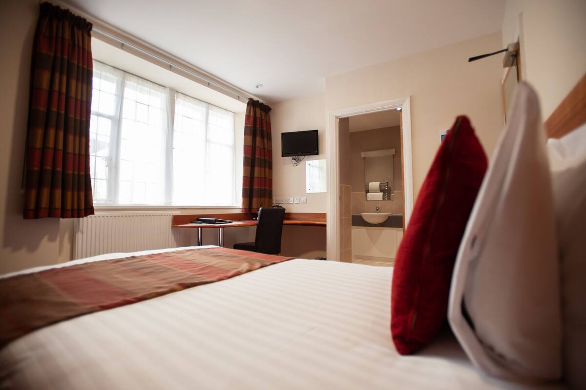 A hotel room with a bed, a desk, a chair, a TV, and a bathroom visible through an open door.