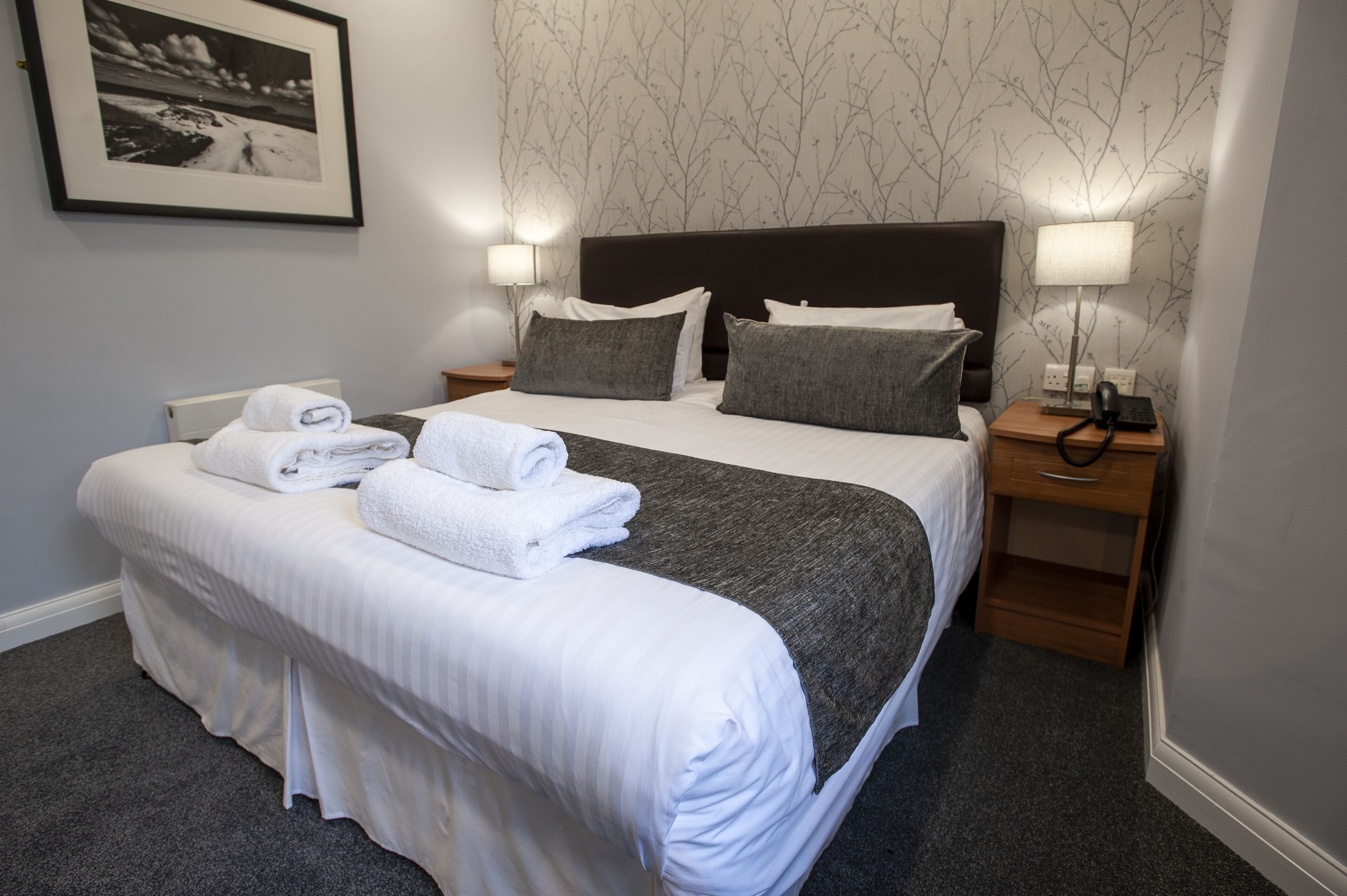 A neatly made bed with white linens and a grey throw blanket, two bedside lamps, a framed picture on the wall, and a small wooden nightstand with a phone and charger.