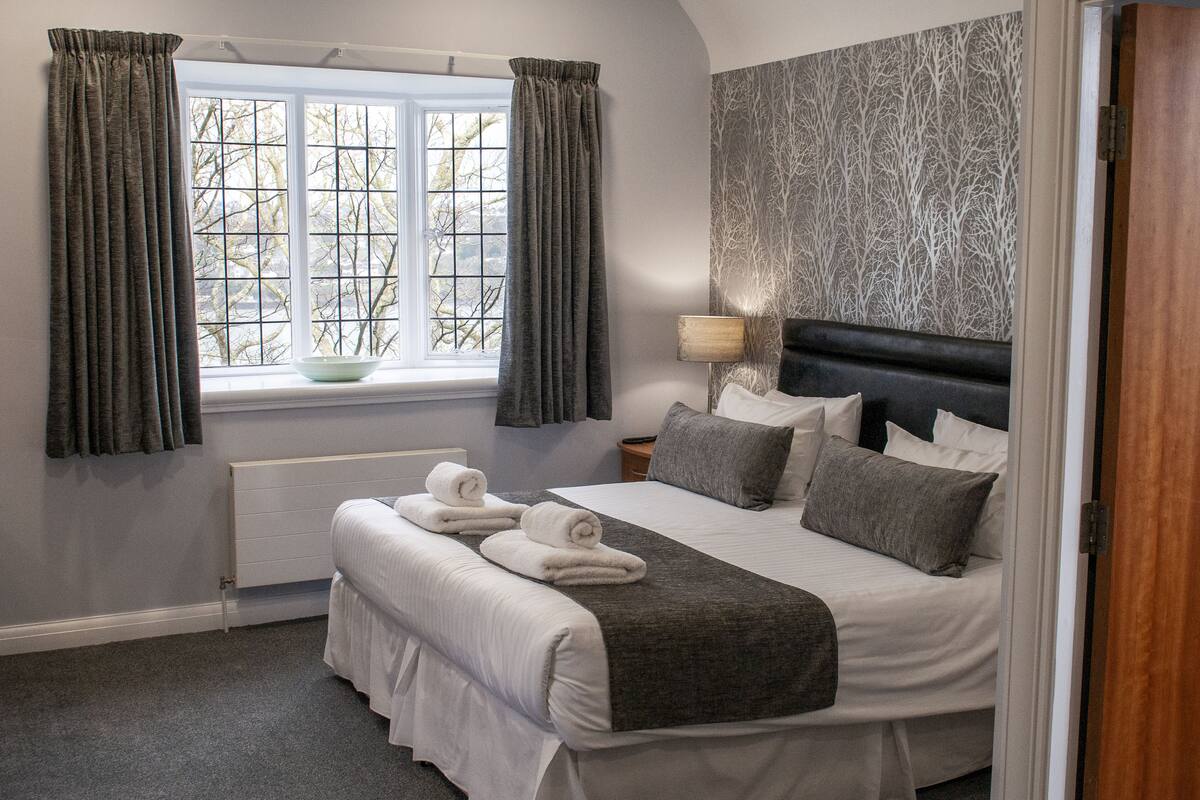 A neatly made bed with pillows and a blanket, a bedside table with a lamp, a window with curtains, and a radiator.