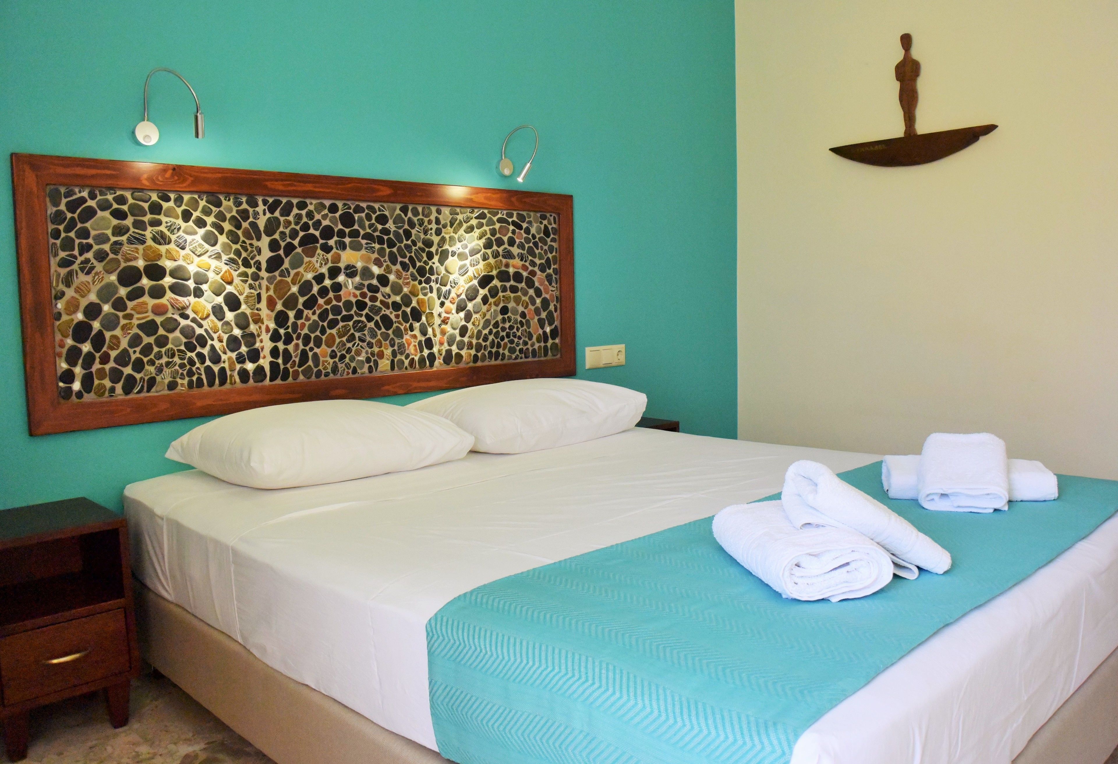 A neatly made bed with white linens and a teal bedspread, a wooden headboard with a mosaic design, and a small nightstand with a drawer.
