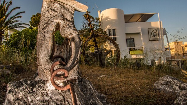 Un pilier de pierre rustique surmonté d’un anneau métallique rouillé, un bâtiment moderne blanc doté d’une section cylindrique et d’un toit plat, le tout entouré de verdure et de palmiers.