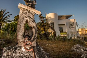 A rustic stone pillar with a rusted metal ring, a modern white building with a cylindrical section and a flat roof, surrounded by greenery and palm trees. - Aris Hotel (Kantanos-Selino)