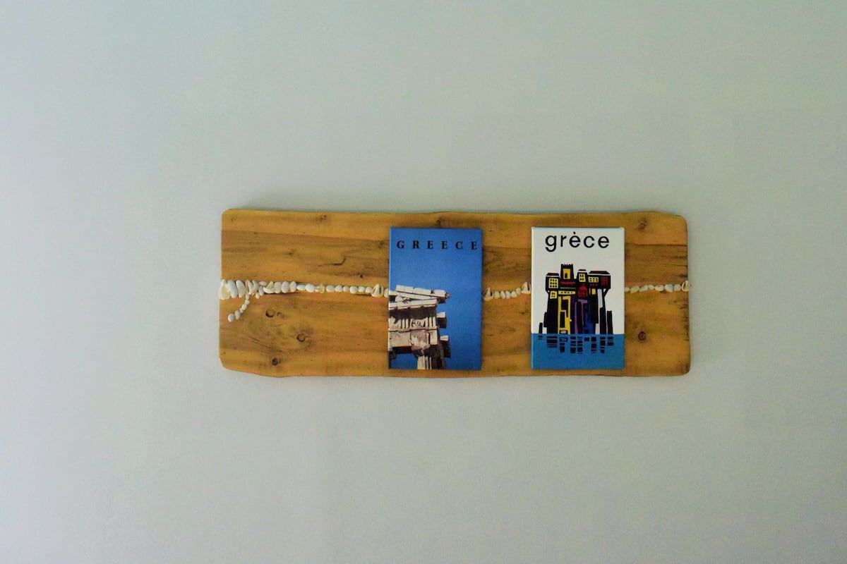 A ceiling fan with three blades and a light fixture, mounted on a light blue wall. Below, a wooden board displays two images with the word "GRECE" and a cityscape illustration.